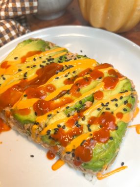 Close-up of an air fryer salmon stack layered with avocado and sushi rice, topped with spicy mayo, Sriracha, and black sesame seeds.