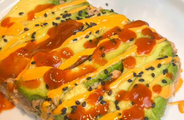 Close-up of an air fryer salmon stack layered with avocado and sushi rice, topped with spicy mayo, Sriracha, and black sesame seeds.