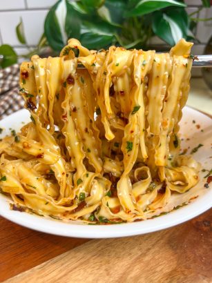 Wide chili crisp noodles lifted over a white bowl, coated with chili oil and herbs