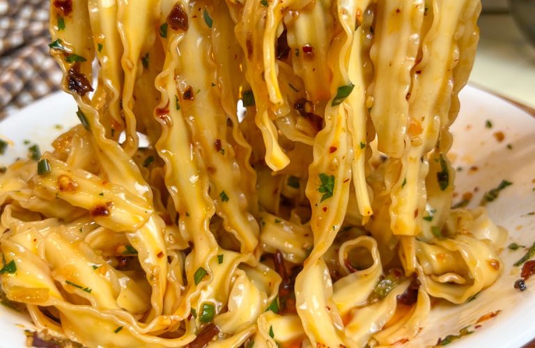 Wide chili crisp noodles lifted over a white bowl, coated with chili oil and herbs